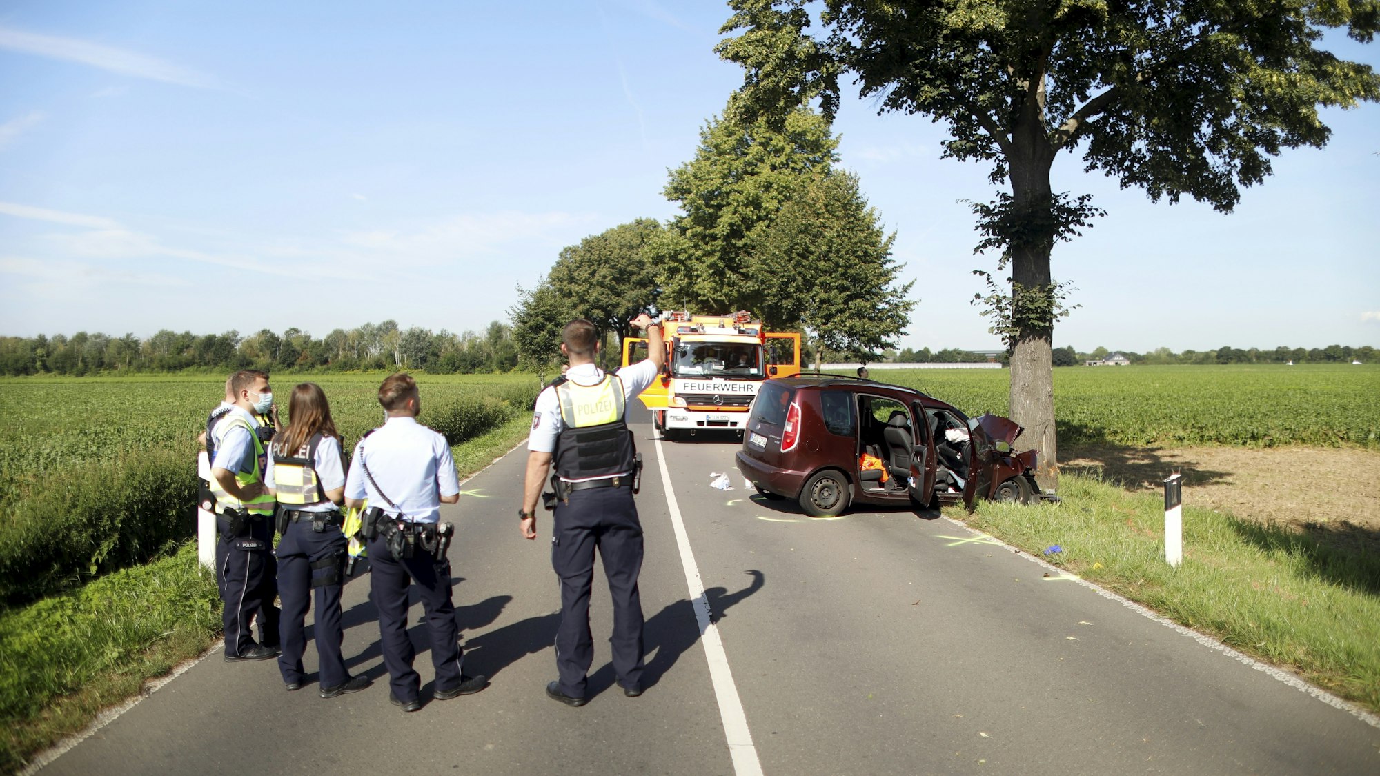 Mehrere Polizisten stehen auf der Fahrbahn und blicken auf das Auto, das schwer beschädigt an einem Straßenbaum steht.