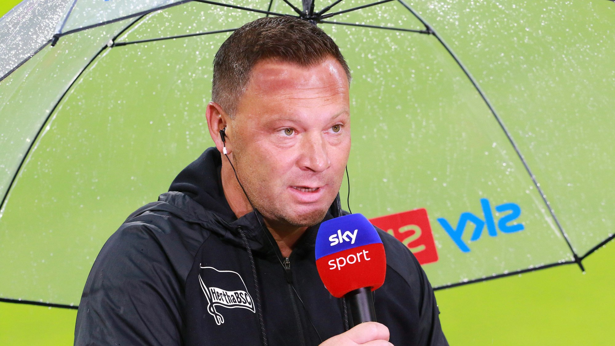 Pal Dardai steht mit einem Mikro unter einem Regenschirm.