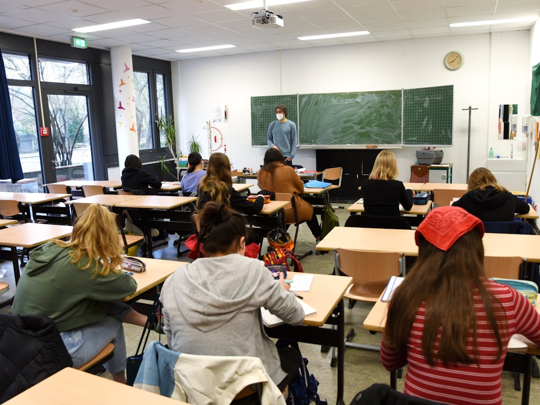 Schüler und Schülerinnen sitzen an ihren Tischen, schreiben und hören dem Lehrer zu.