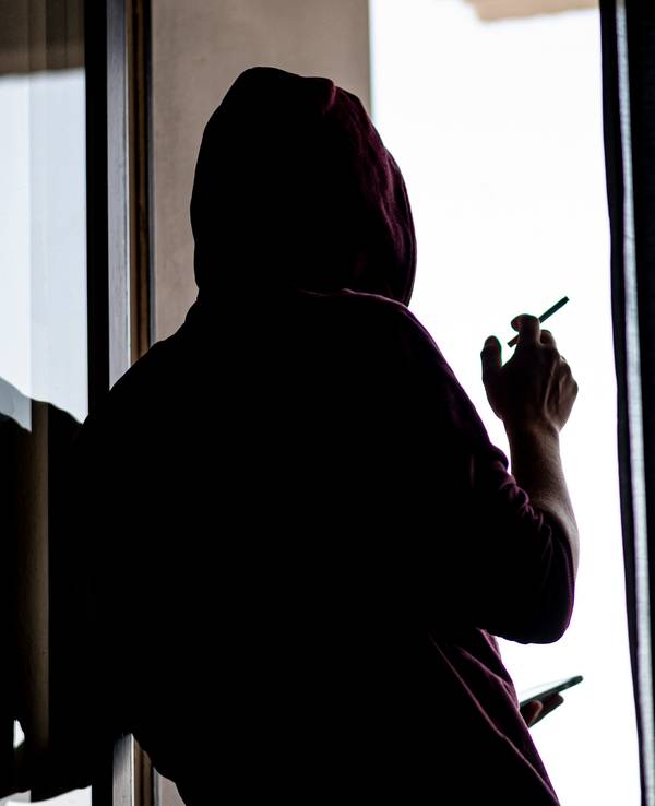 Ein junger Mann steht mit Zigarette und Smartphone an einem Fenster.