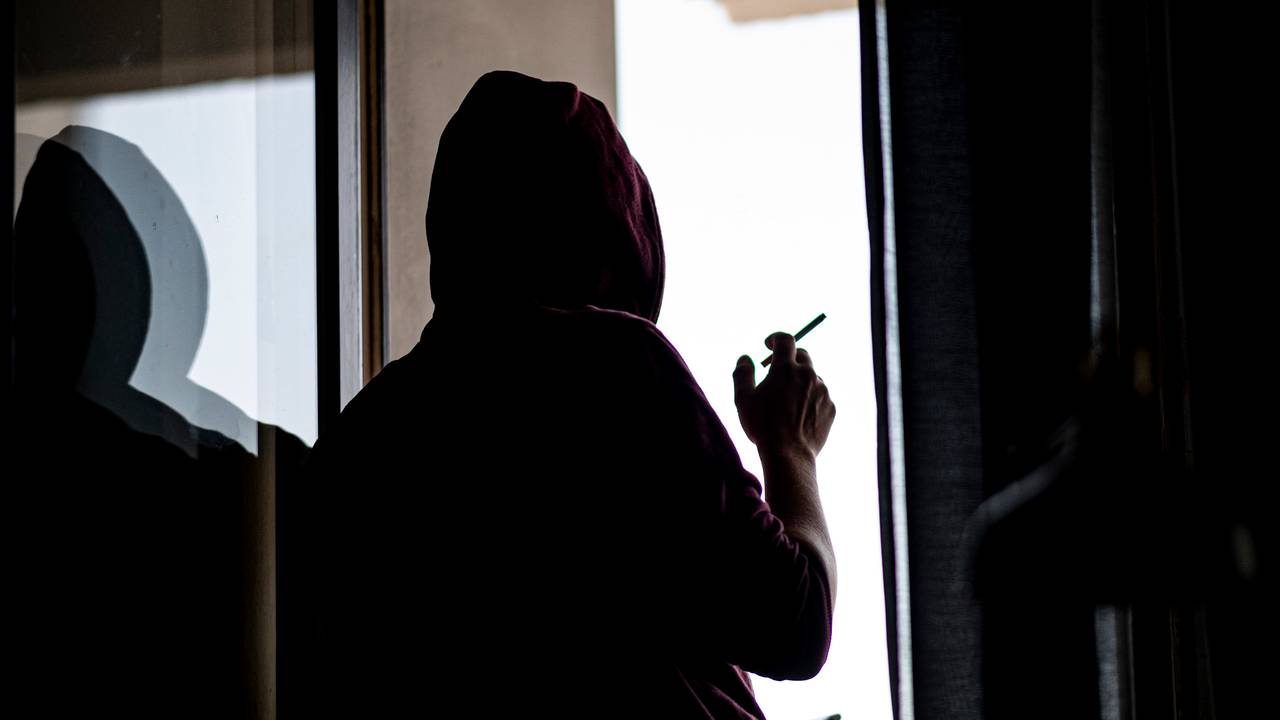 Ein junger Mann steht mit Zigarette und Smartphone an einem Fenster.