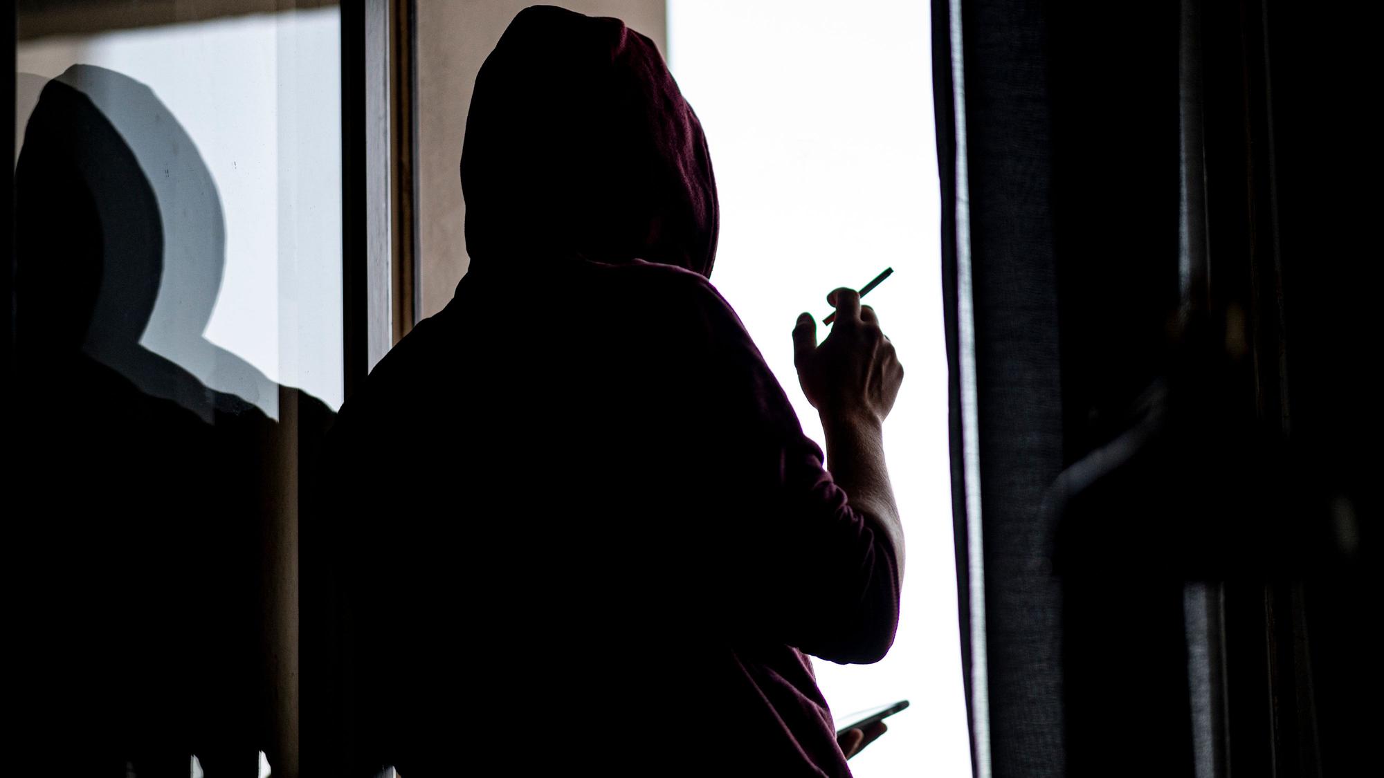 Ein junger Mann steht mit Zigarette und Smartphone an einem Fenster.