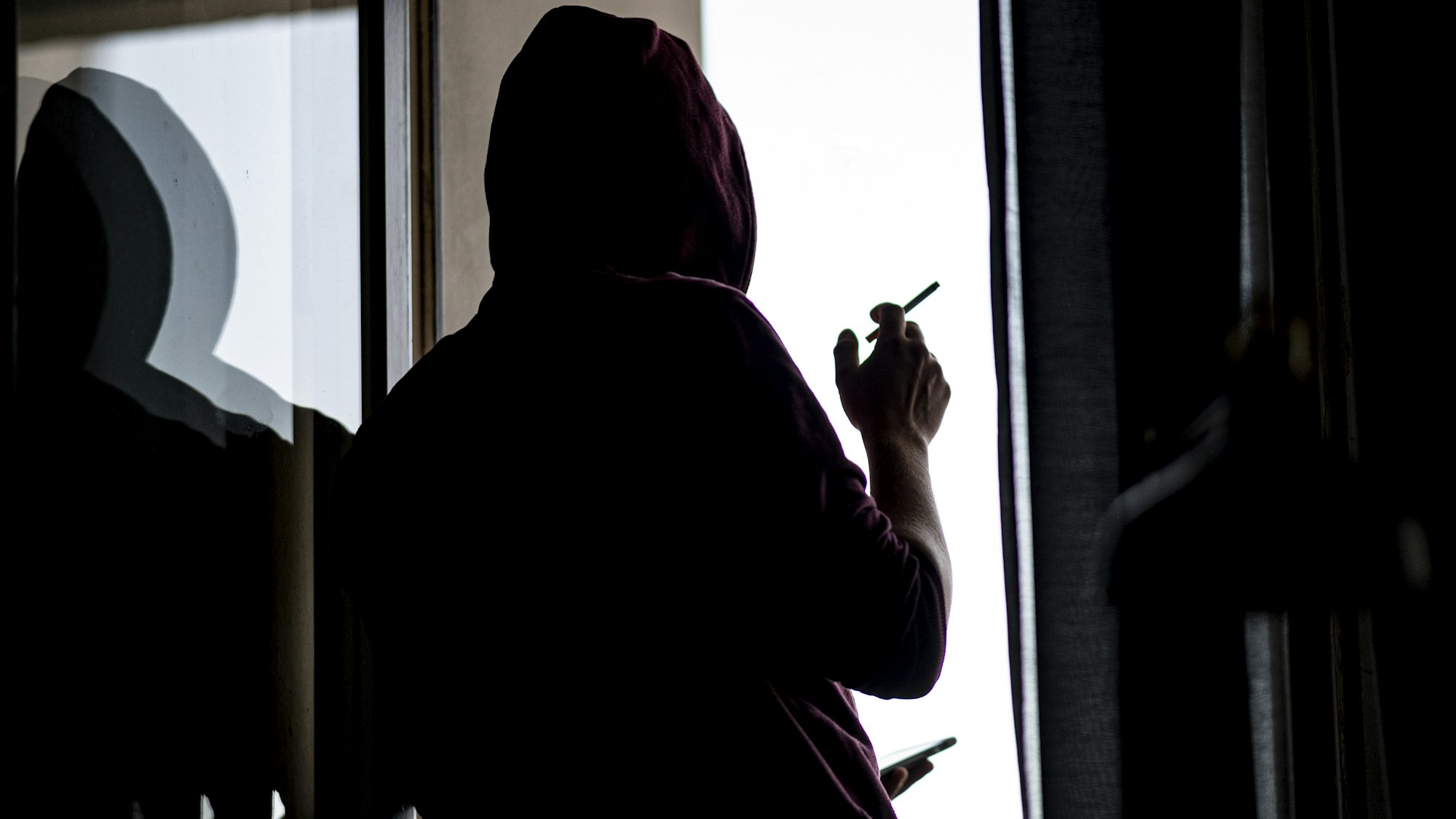 Ein junger Mann steht mit Zigarette und Smartphone an einem Fenster.