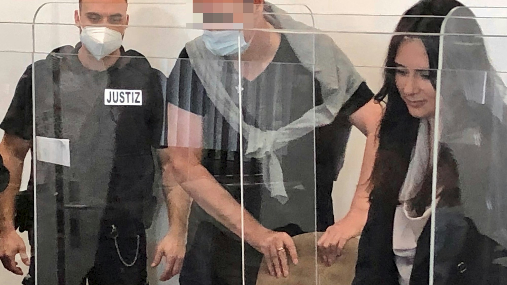 Der Angeklagte hat die Lehne eines Stuhls gepackt, rechts steht seine Anwältin, links ein Justizvollzugsbeamter.