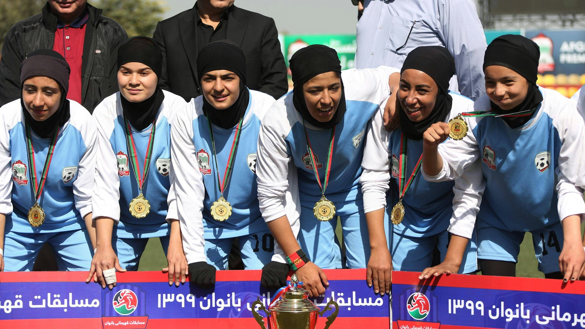 Sechs Spielerinnen von Herat sind zu sehen. Sie tragen Trikots, Kopftücher und Medaillen. Vor ihnen steht der Pokal.