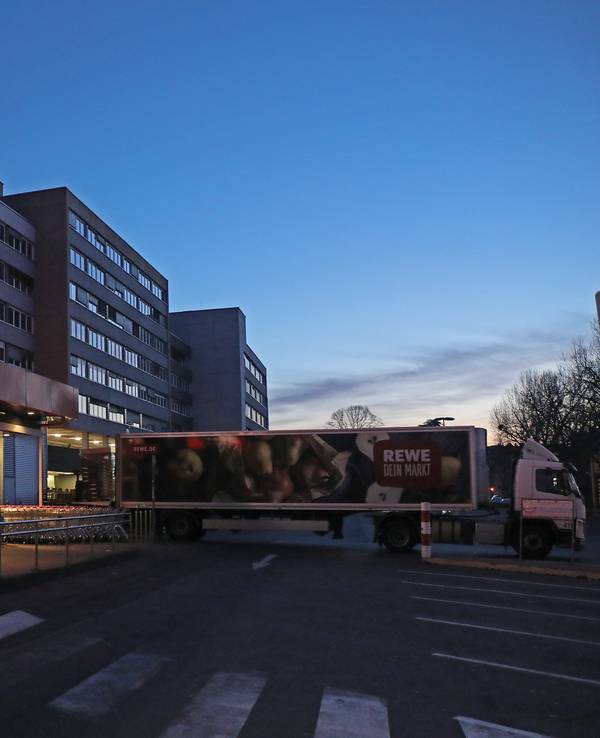 Ein Lkw steht am frühen Morgen vor einem Rewe Supermarkt, der mit Waren beliefert wird. Es ist dunkel, im Hintergrund ist ein Hochhaus zu sehen. Es ist fast menschenleer.