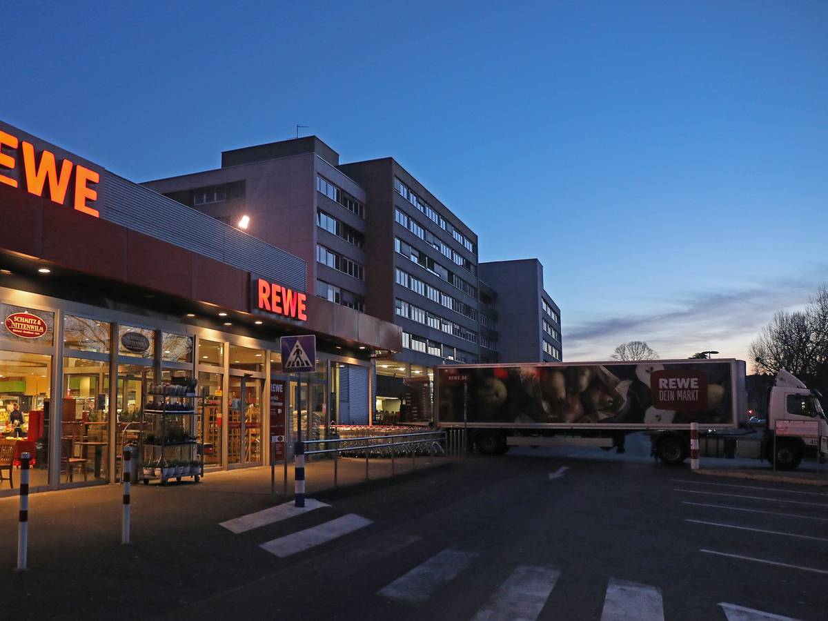 Ein Lkw steht am frühen Morgen vor einem Rewe Supermarkt, der mit Waren beliefert wird. Es ist dunkel, im Hintergrund ist ein Hochhaus zu sehen. Es ist fast menschenleer.