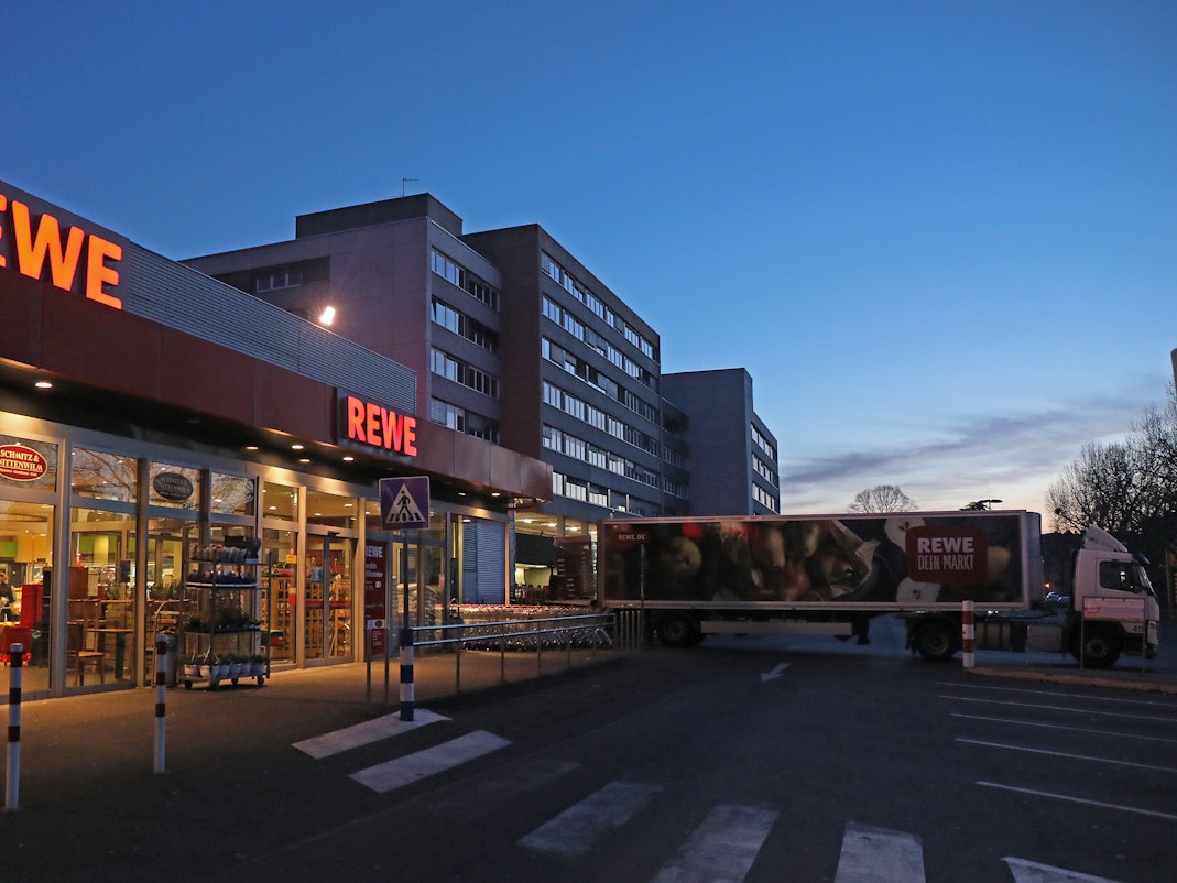 Ein Lkw steht am frühen Morgen vor einem Rewe Supermarkt, der mit Waren beliefert wird. Es ist dunkel, im Hintergrund ist ein Hochhaus zu sehen. Es ist fast menschenleer.