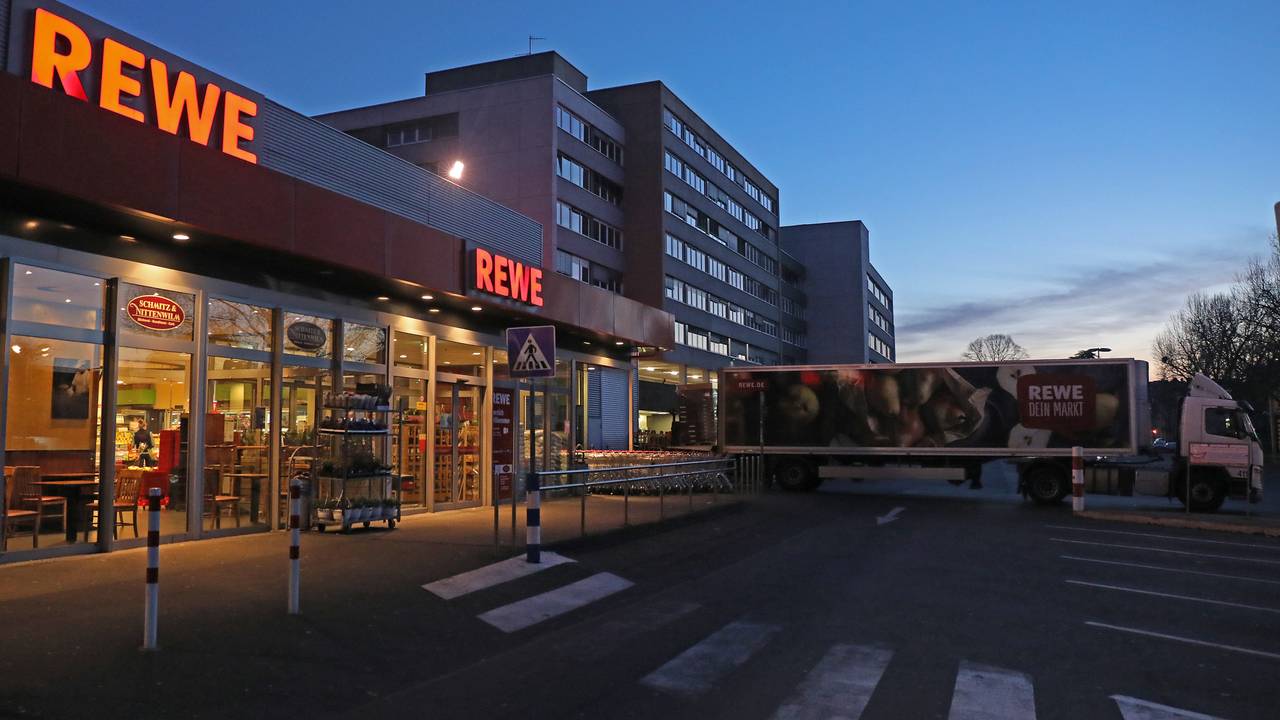 Ein Lkw steht am frühen Morgen vor einem Rewe Supermarkt, der mit Waren beliefert wird. Es ist dunkel, im Hintergrund ist ein Hochhaus zu sehen. Es ist fast menschenleer.