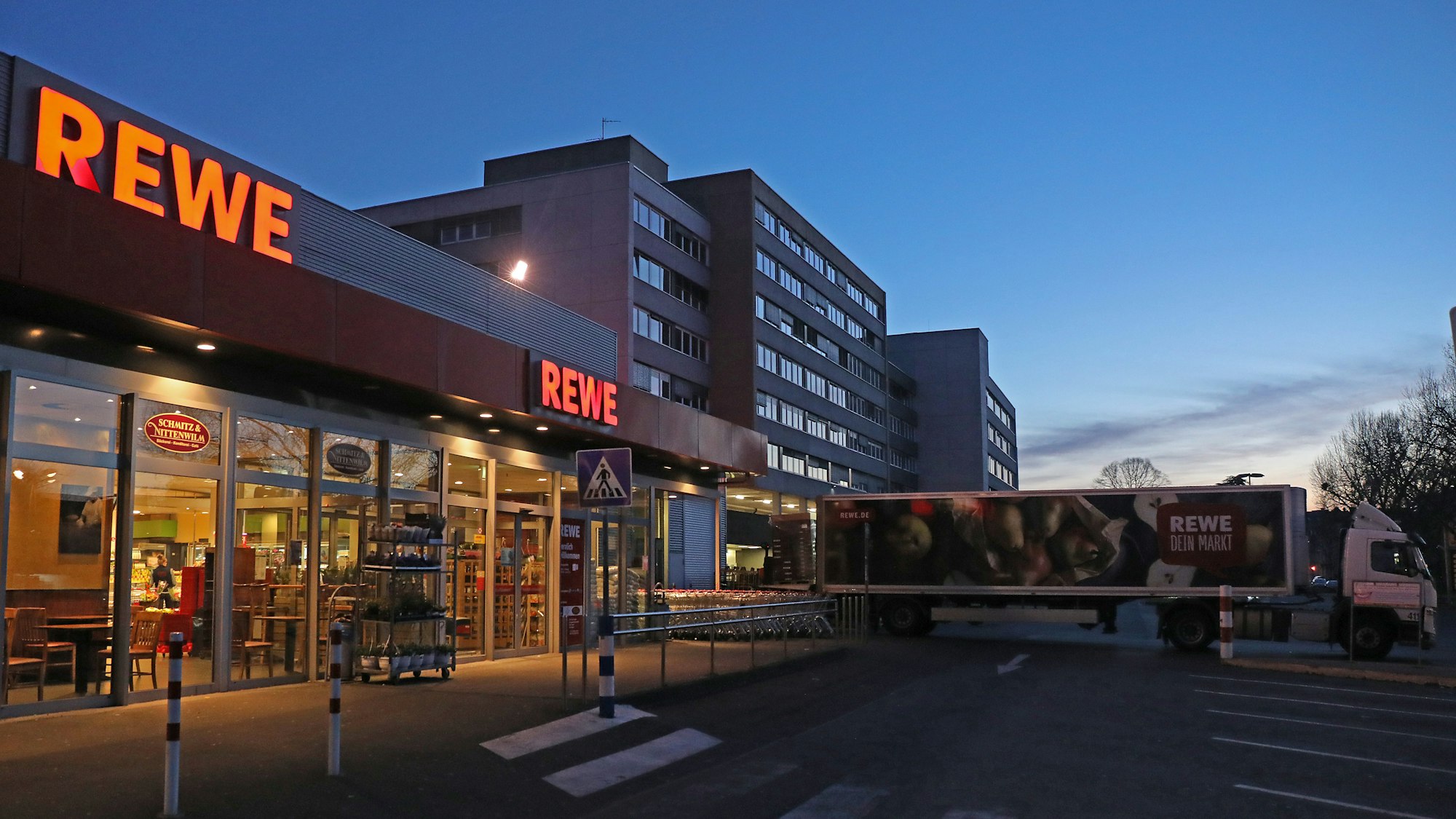 Ein Lkw steht am frühen Morgen vor einem Rewe Supermarkt, der mit Waren beliefert wird. Es ist dunkel, im Hintergrund ist ein Hochhaus zu sehen. Es ist fast menschenleer.