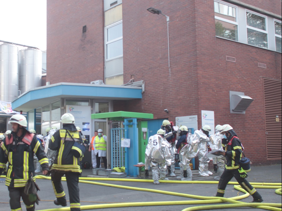 Feuerwehrmänner und Männer in Spezialanzügen befinden sich vor einem Werk.