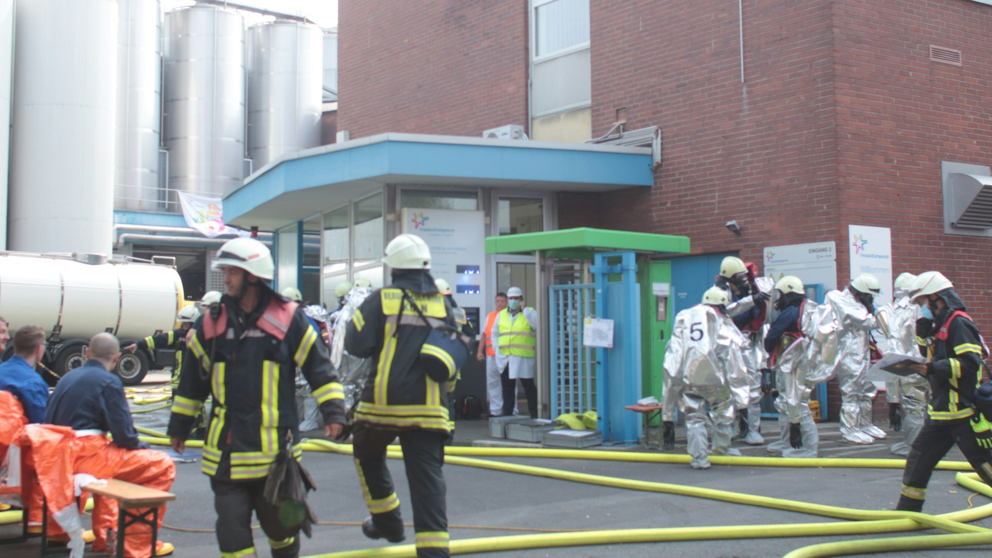 Feuerwehrmänner und Männer in Spezialanzügen befinden sich vor einem Werk.