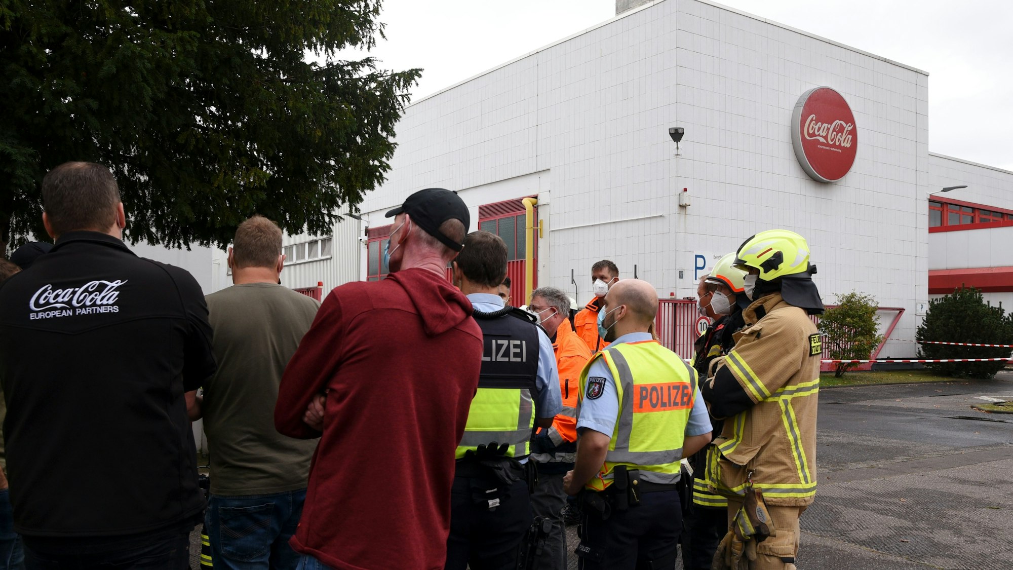 Polizei, Feuerwehr und Coca-Cola-Mitarbeiter stehen vor dem Werk in Köln-Ossendorf.