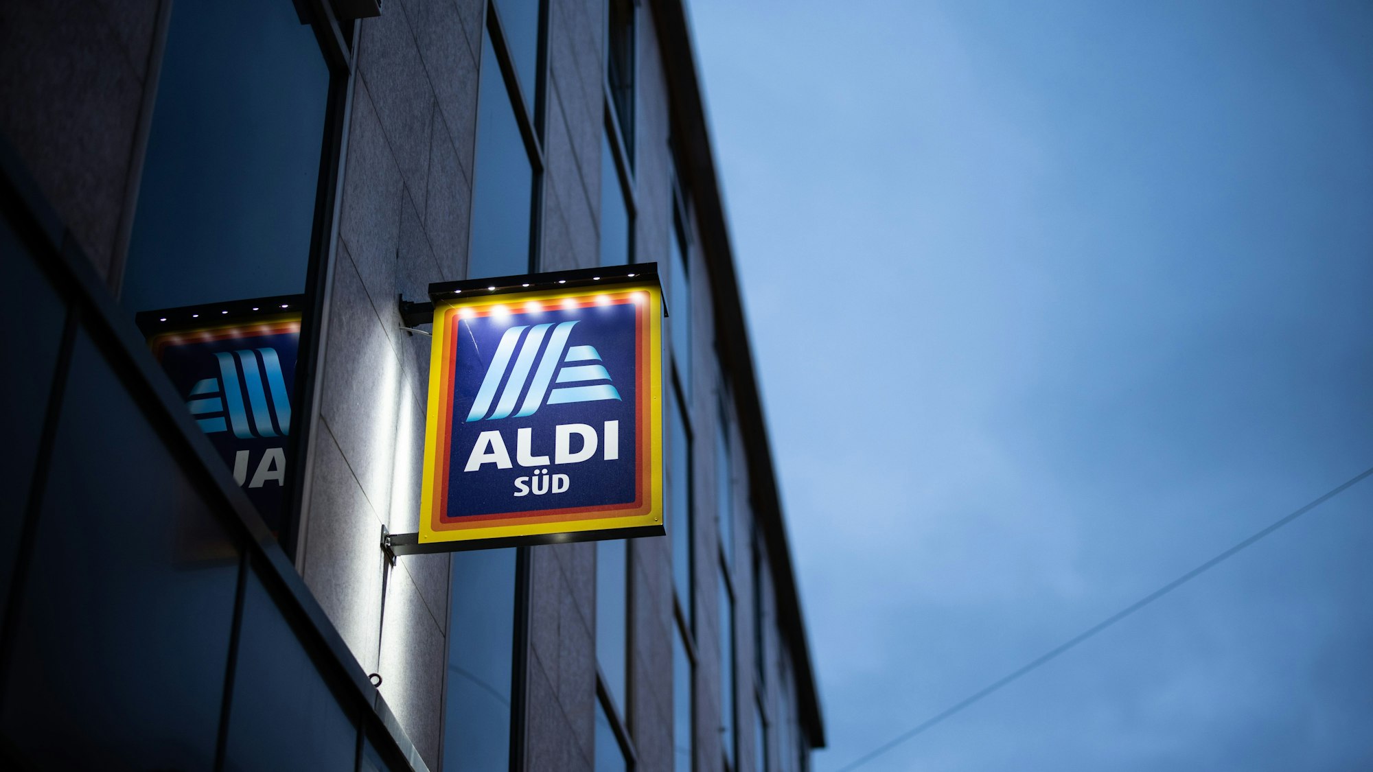 Das Logo einer Aldi Süd Filiale, fotografiert in einer Fußgängerzone der Düsseldorfer Innenstadt, ist beleuchtet am Abend zu sehen.