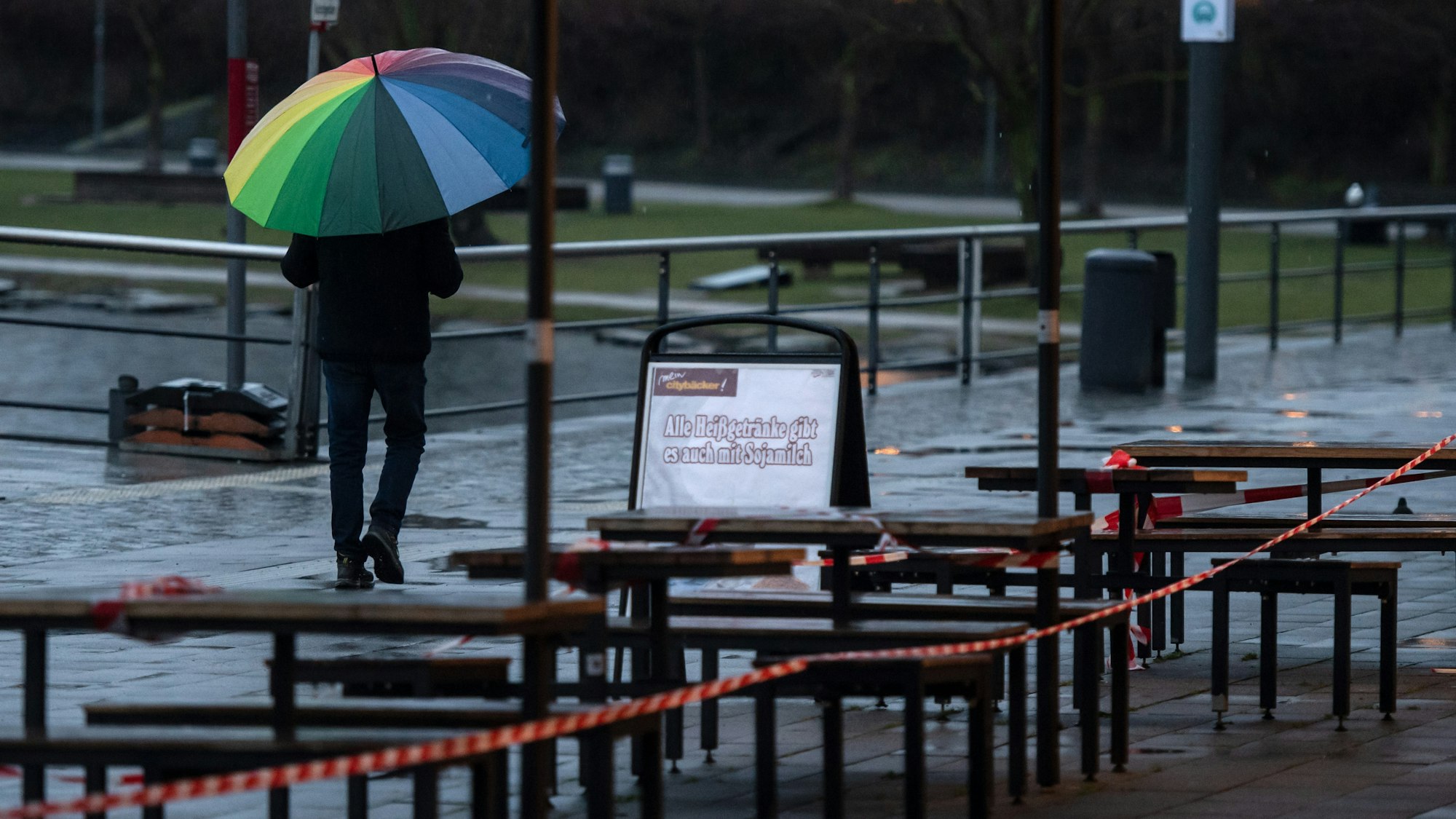 Ein Passant geht unter einem bunten Regenschirm an der Uferprommenade des Phoenix Sees an den wegen der Corona-Pandemie gesperrten Tischen vor einer Bäckerei vorbei.