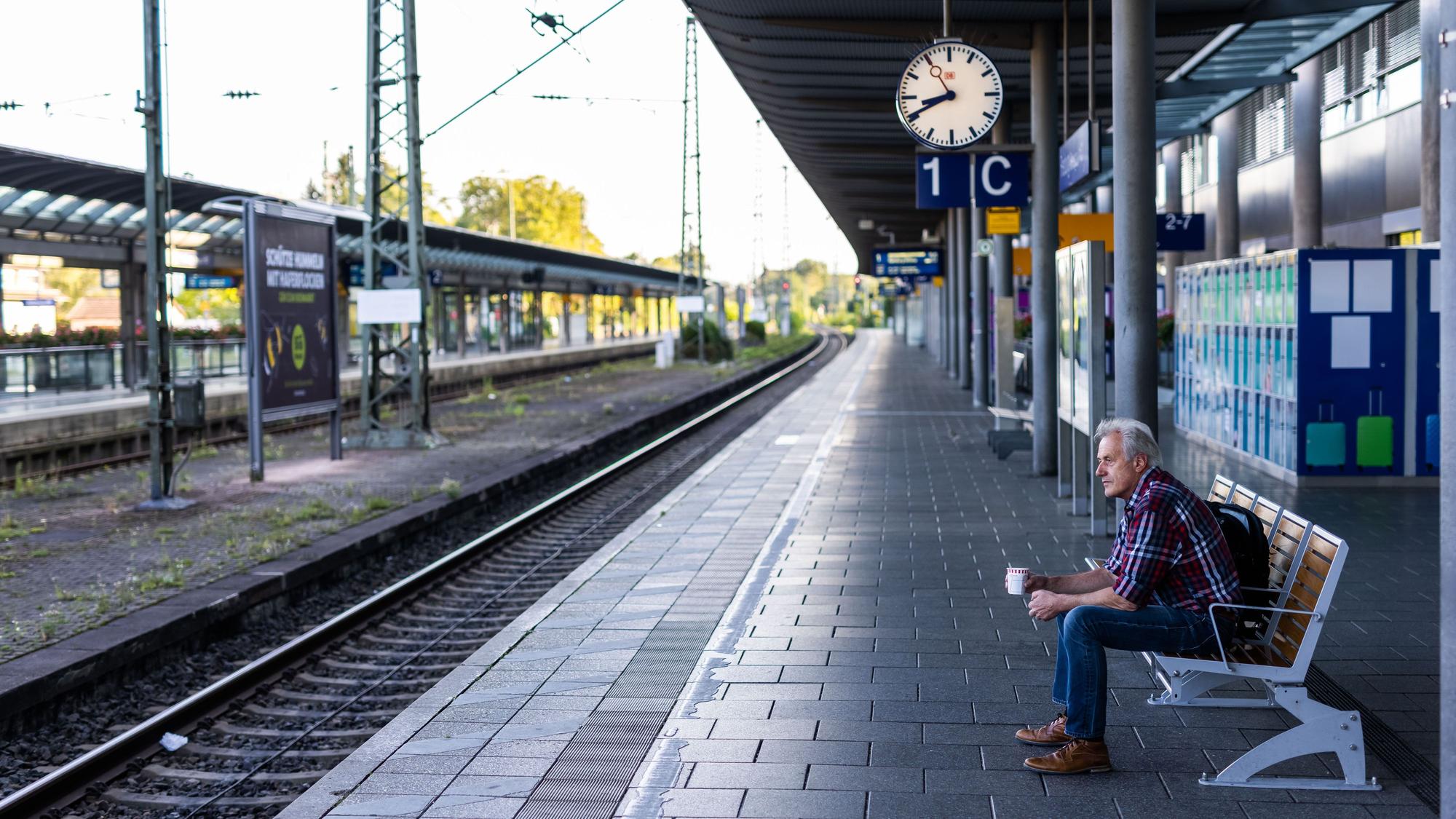 Ein Mann sitzt mit einem Kaffee in der Hand auf einer Bank auf dem ansonsten menschenleeren Bahnsteig.