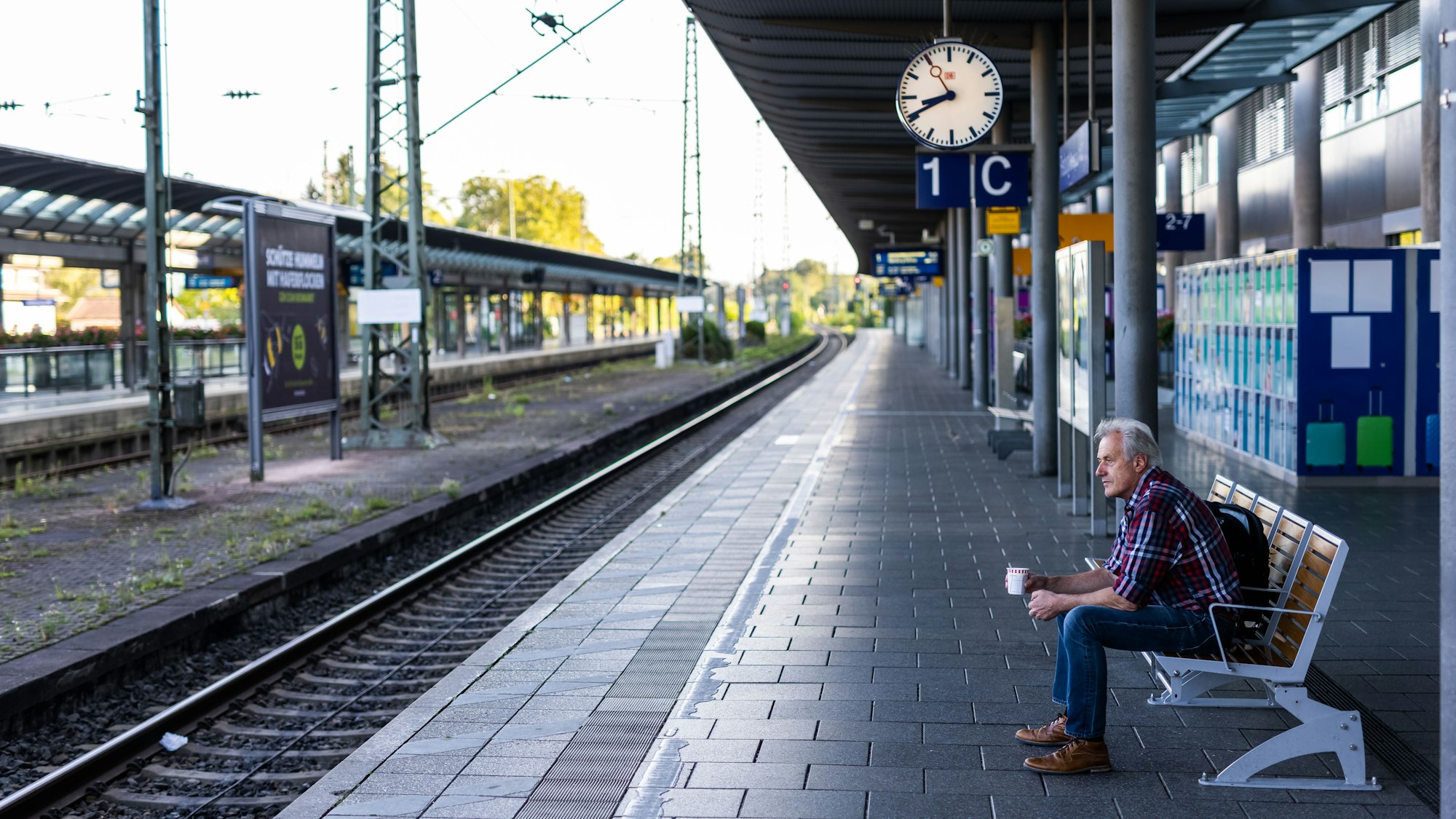 Ein Mann sitzt mit einem Kaffee in der Hand auf einer Bank auf dem ansonsten menschenleeren Bahnsteig.