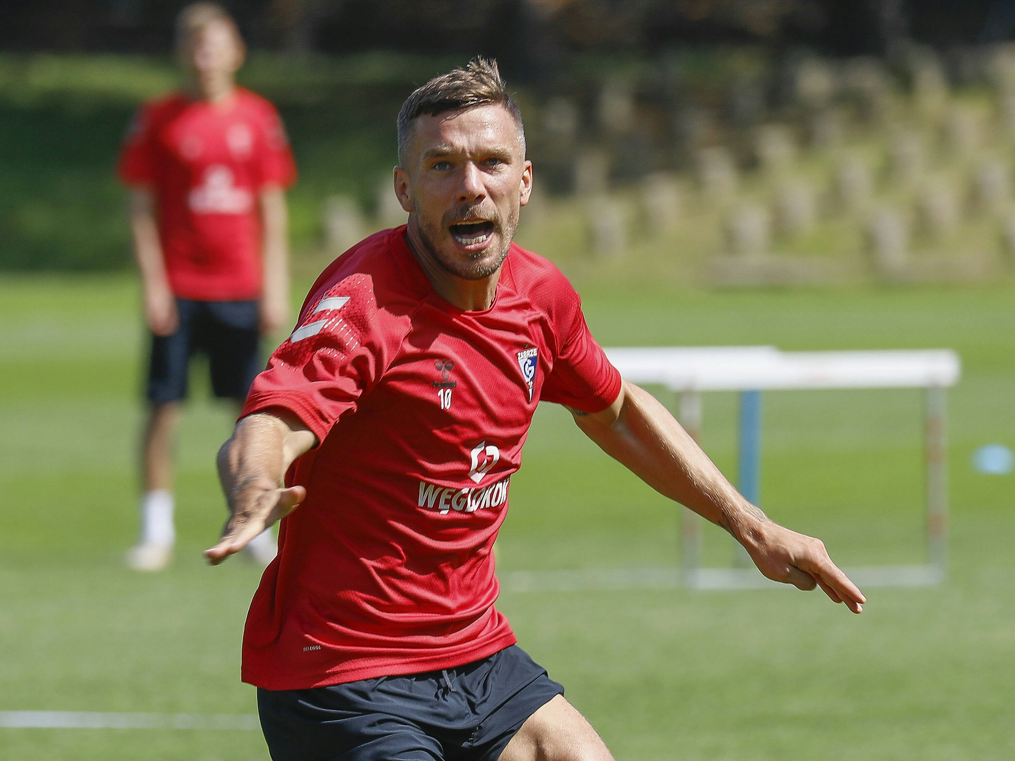 Podolski reißt auf dem Trainingsplatz den Mund auf und breitet die Arme auf.