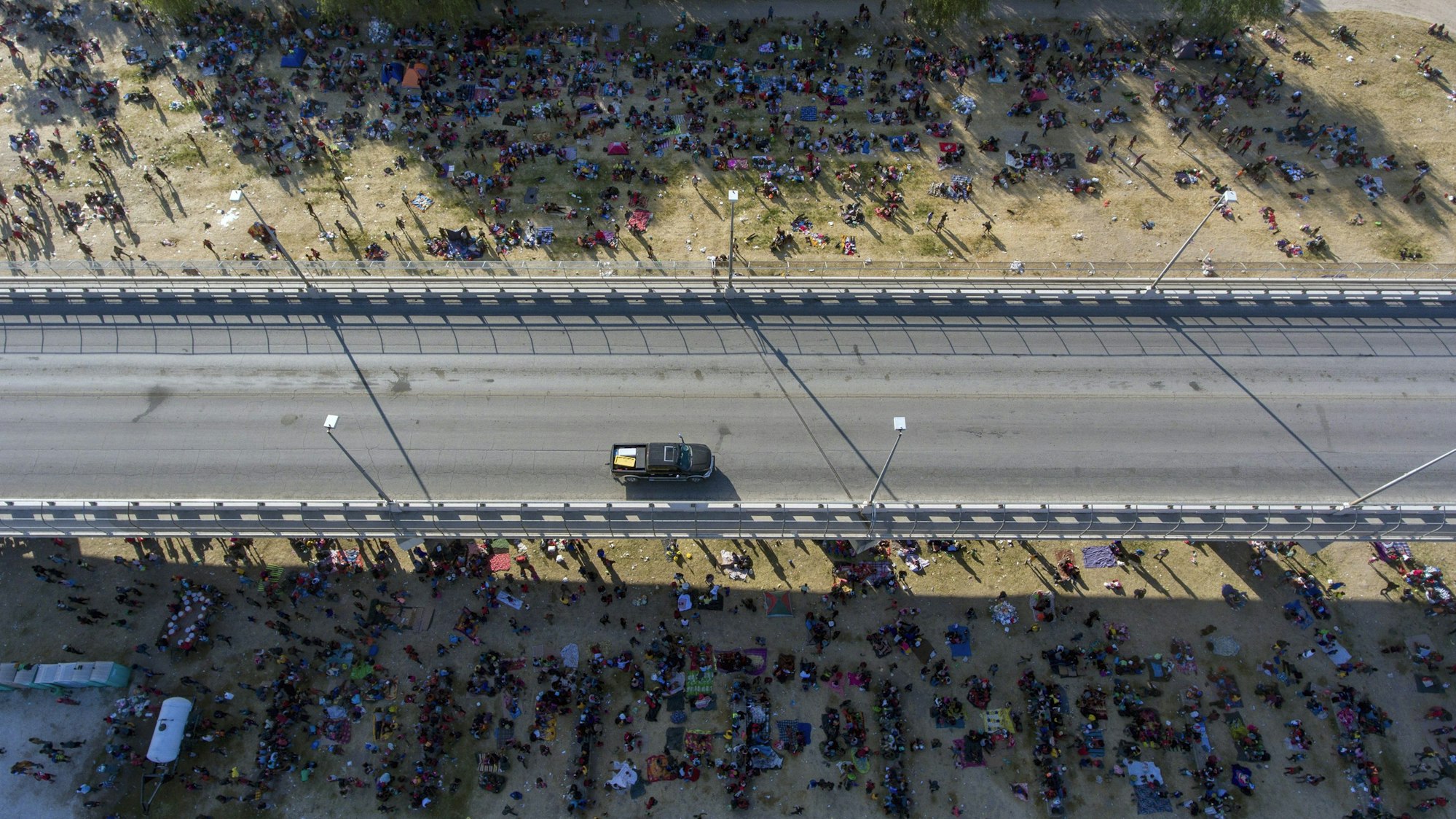 Migranten kampieren unter einer Brücke in Del Rio. Eine Straße ist von oben zu sehen, an der Seite sind unzählige Menschen zu sehen, die dort im Schatten der Brücke verbleiben.