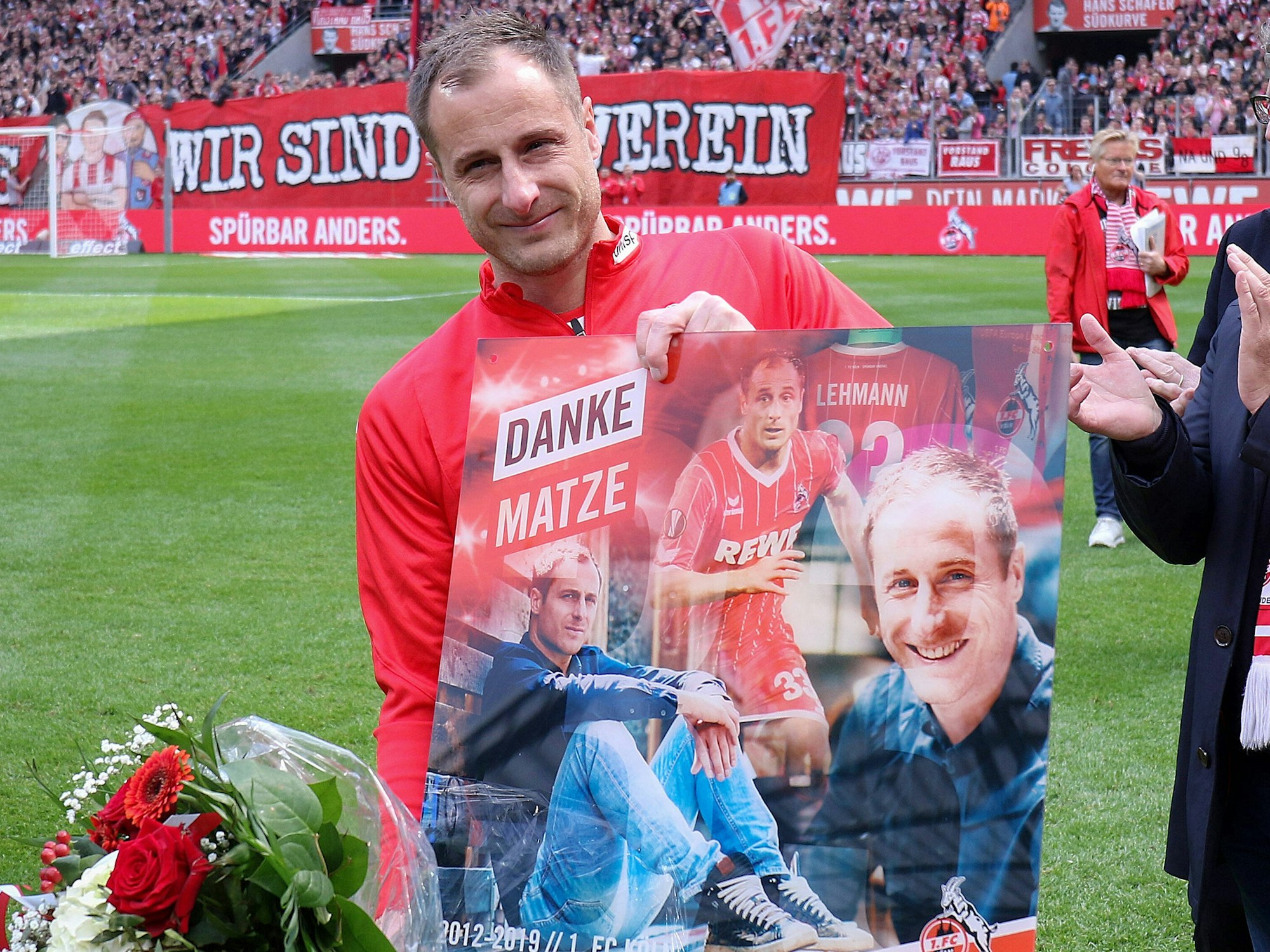 Matthias Lehmann hält ein Bild hoch, auf dem er mehrfach abgebildet ist. Er hält außerdem einen Blumenstrauß in der Hand.