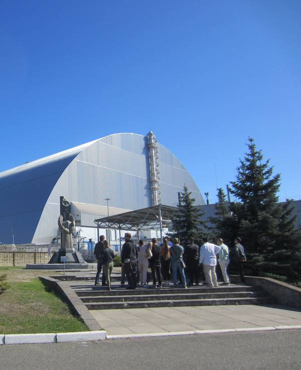 Touristengruppe am Mahnmal vor dem 1986 explodierten Blocks vier des Atomkraftwerks Tschernobyl in der Ukraine. Vor dem geschützten Block steht eine Statue, davor steht eine Gruppe von Touristen.