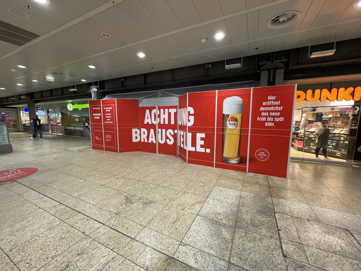 Ein Bauschild mit der Aufschrift „Achtung Braustelle“ steht im Kölner Hauptbahnhof und kündigt das neue Lokal der Brauerei Früh an.