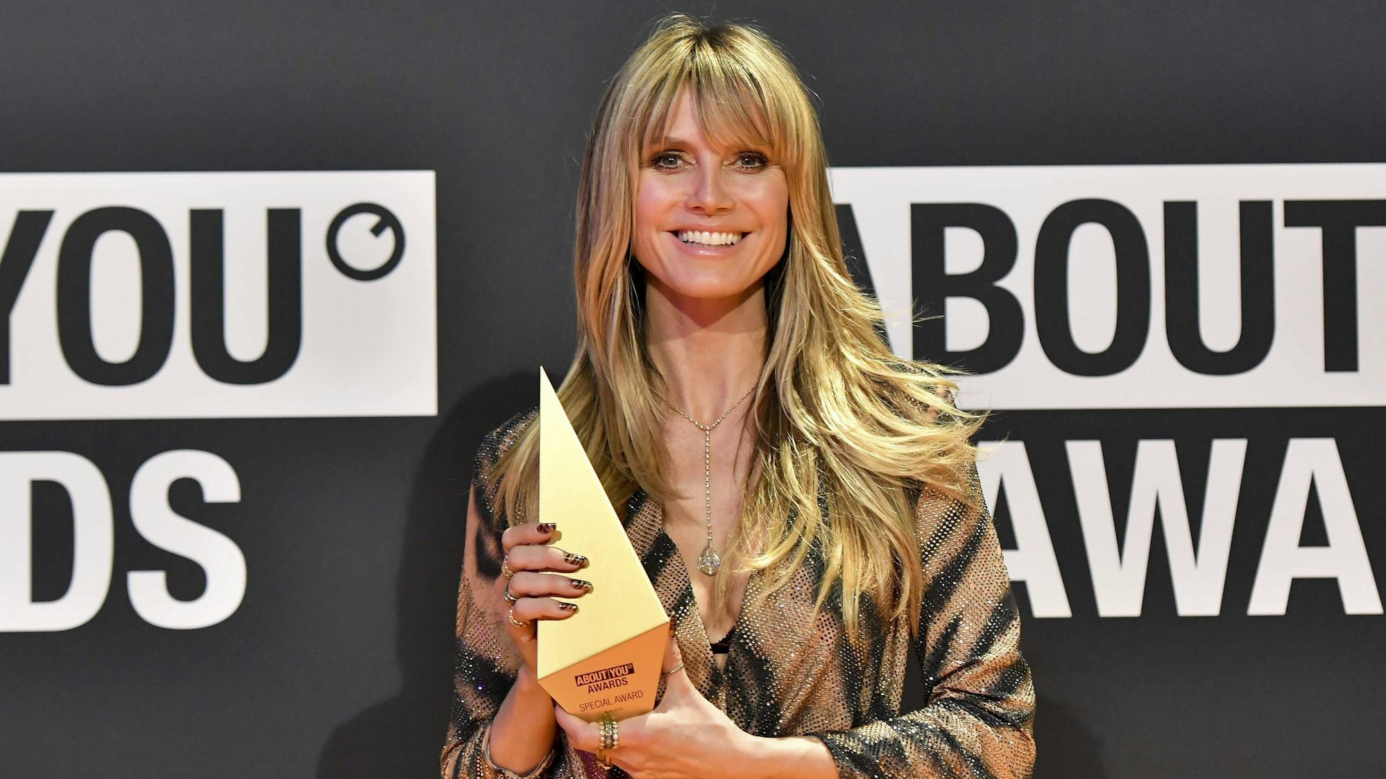 Heidi Klum lächelt mit einem ABOU YOU Trophäe in der Hand in die Kamera.