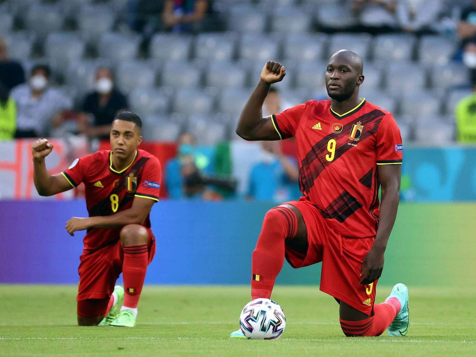 Youri Tielemans und Romelu Lukaku knien am Mittelkreis und heben die rechte Faust.