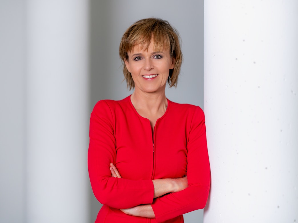 Katja Horneffer steht mit verschränkten Armen an eine Säule gelehnt und lächelt.