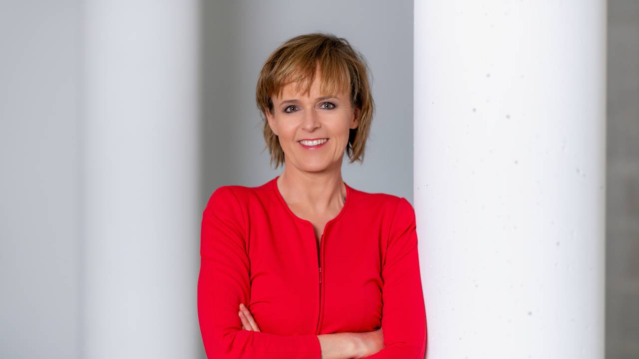 Katja Horneffer steht mit verschränkten Armen an eine Säule gelehnt und lächelt.