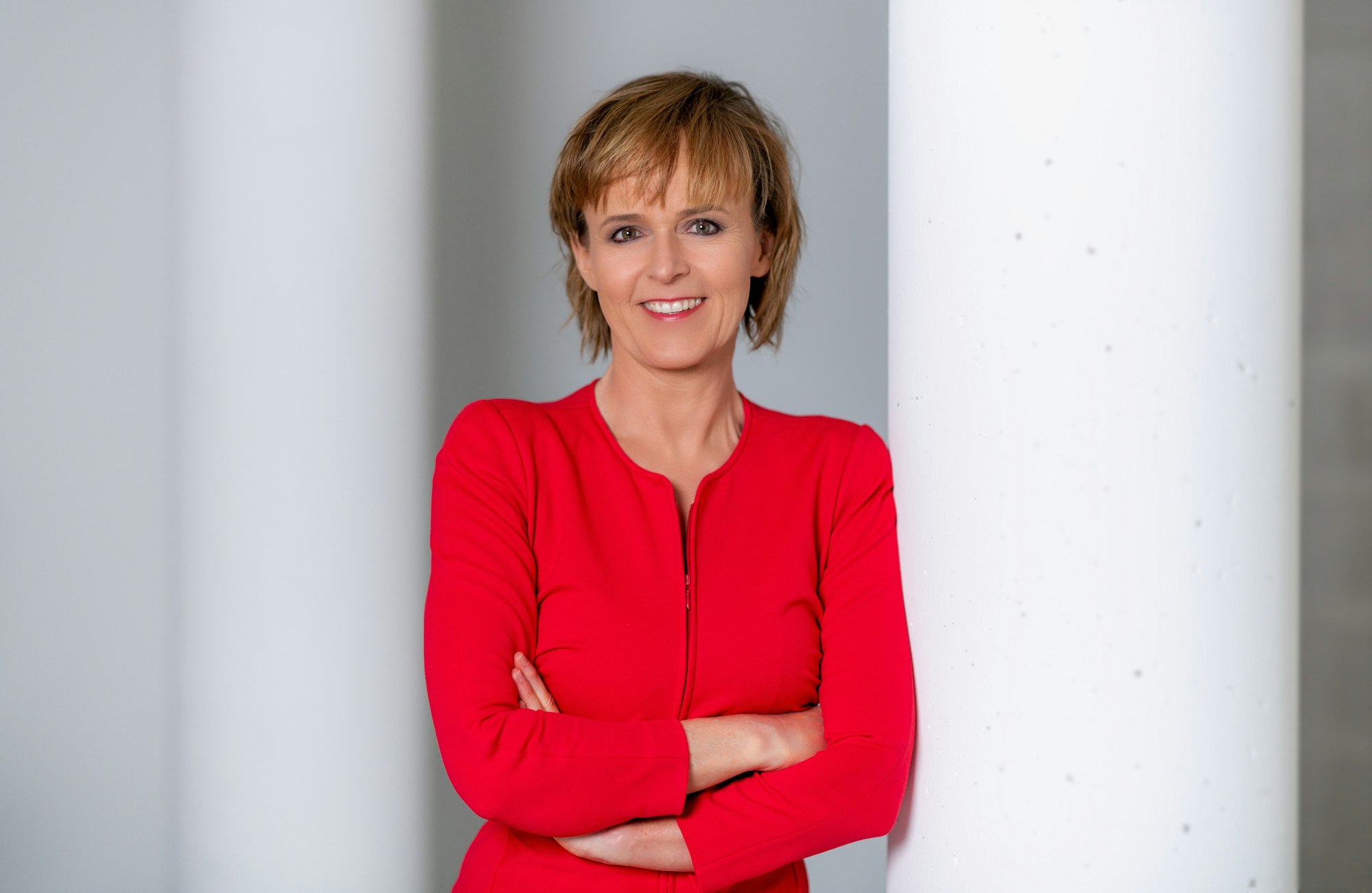 Katja Horneffer steht mit verschränkten Armen an eine Säule gelehnt und lächelt.