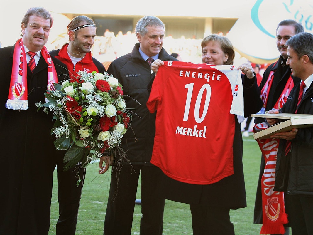 Angela Merkel hält ein Cottbus-Trikot mit der Nummer 10 hoch, Tomislav Piplica hält einen Blumenstrauß in den Händen.