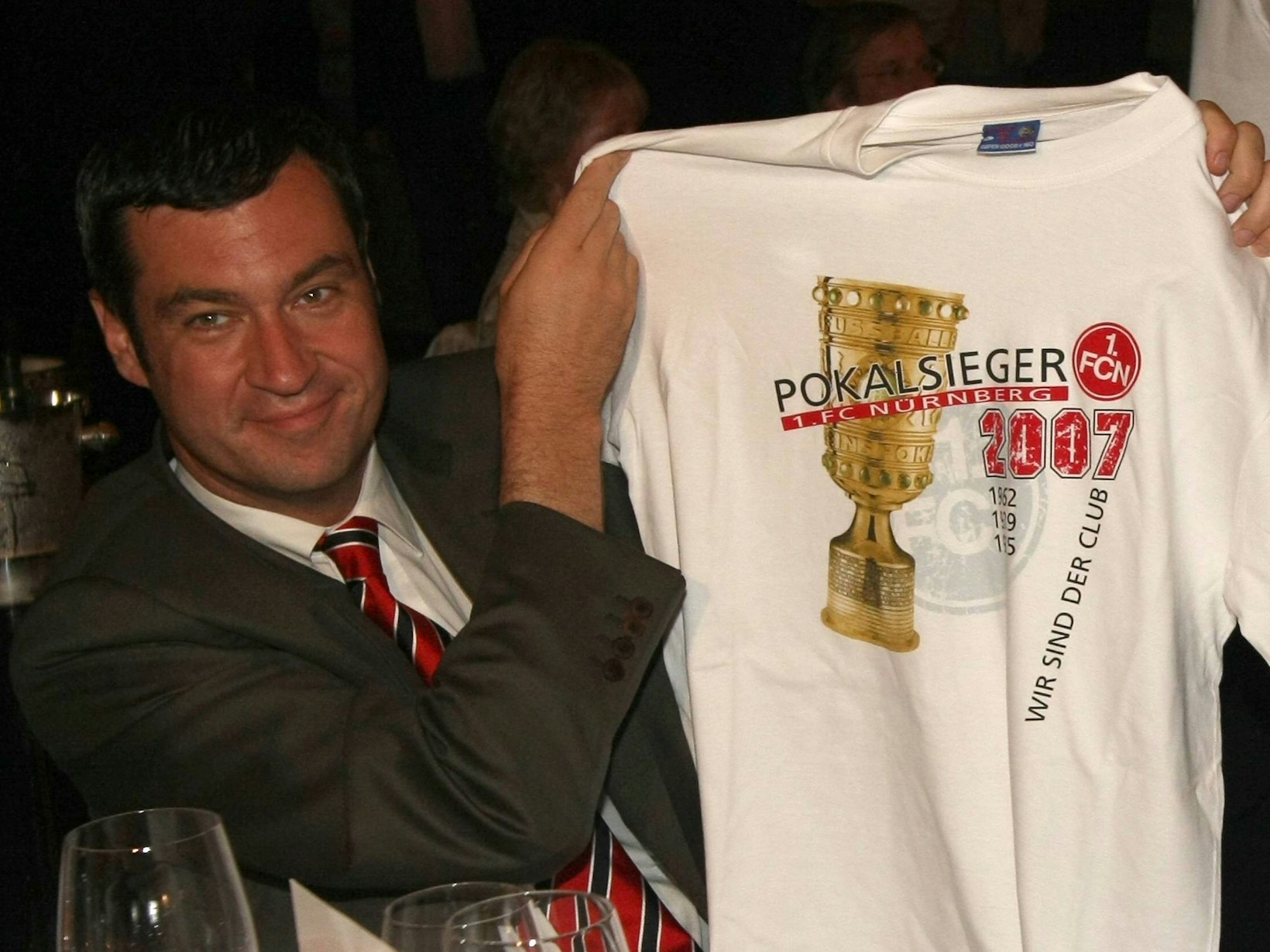 Markus Söder hält ein weißes T-Shirt hoch, auf dem Pokalsieger 2007 steht und der DFB-Pokal abgebildet ist.