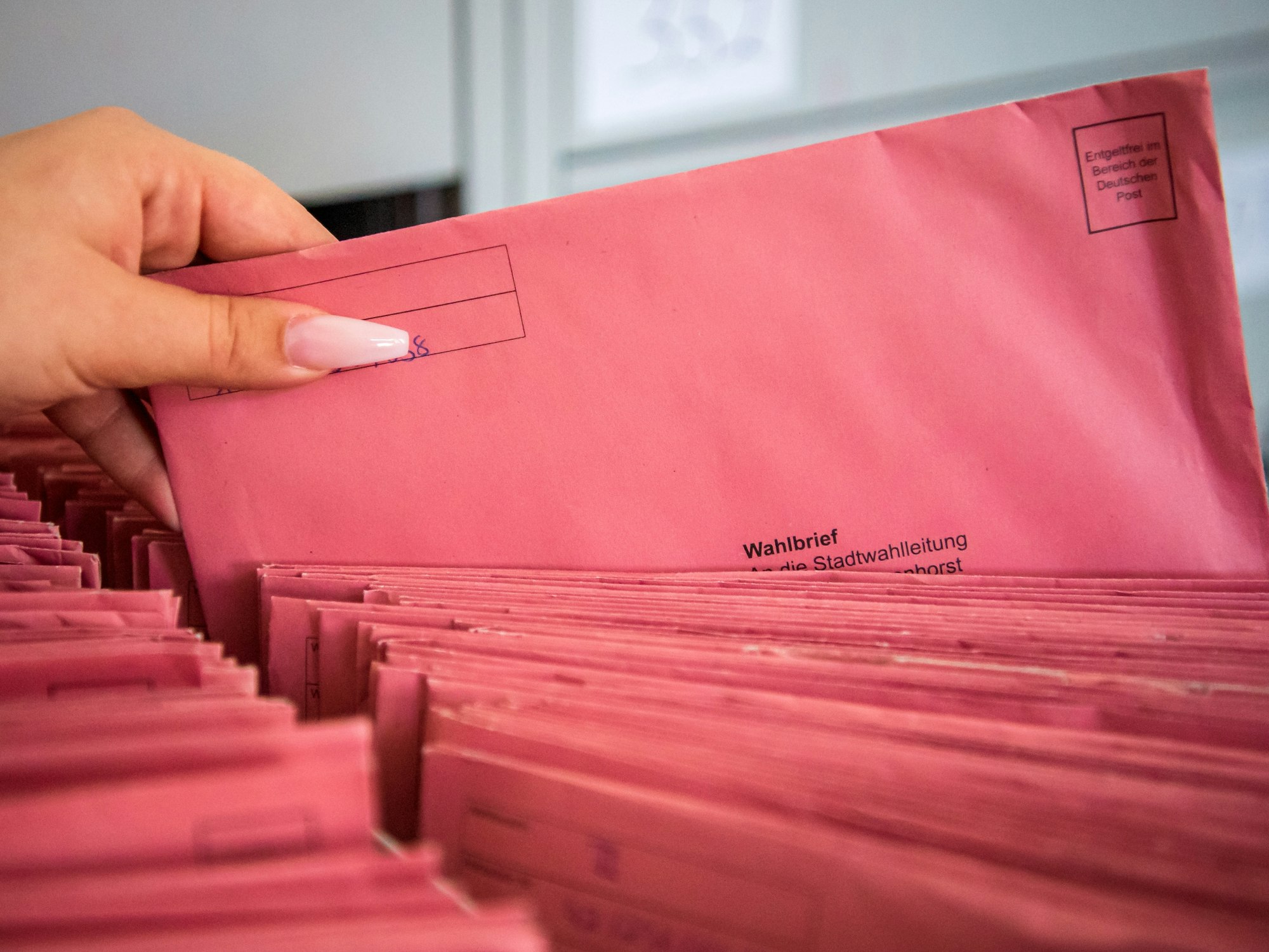 Eine Frauenhand zieht einen Wahlbrief aus einer Kiste, in der sich zahlreiche identische Wahlbriefe befinden.