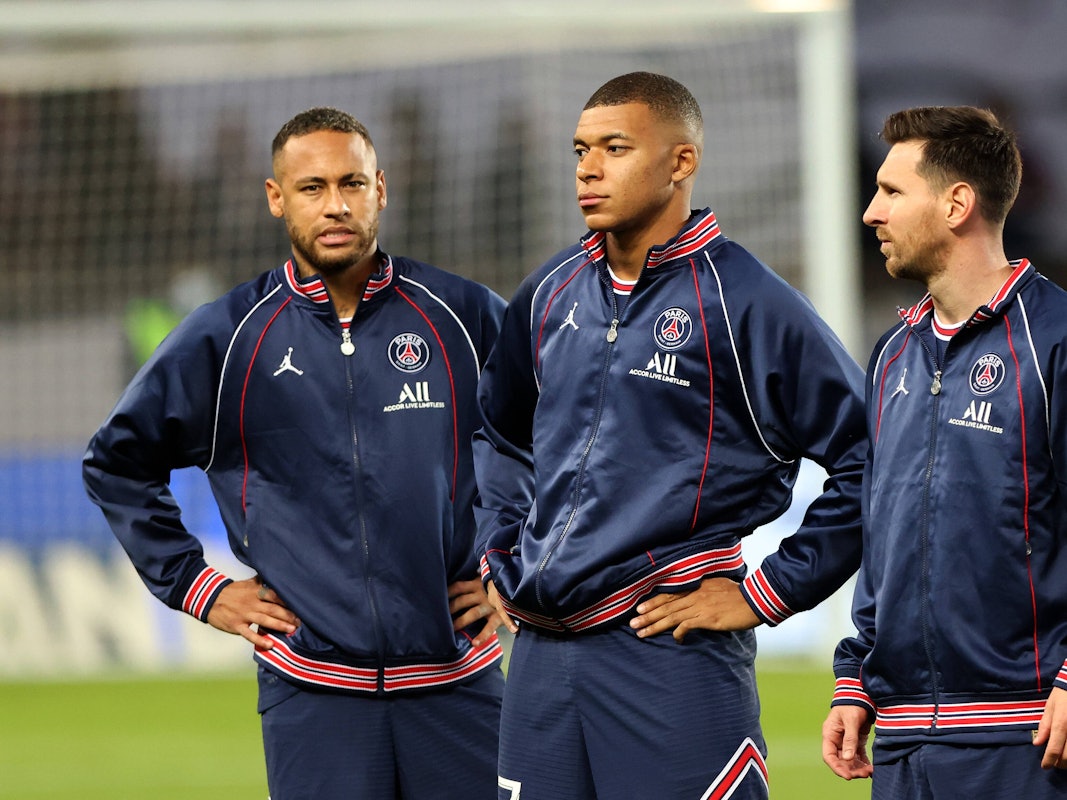 Neymar, Kylian Mbappe und Lionel Messi stehen in Trainingsjacken auf dem Feld. Neymar und Mbappe haben die Hände in den Hüften.