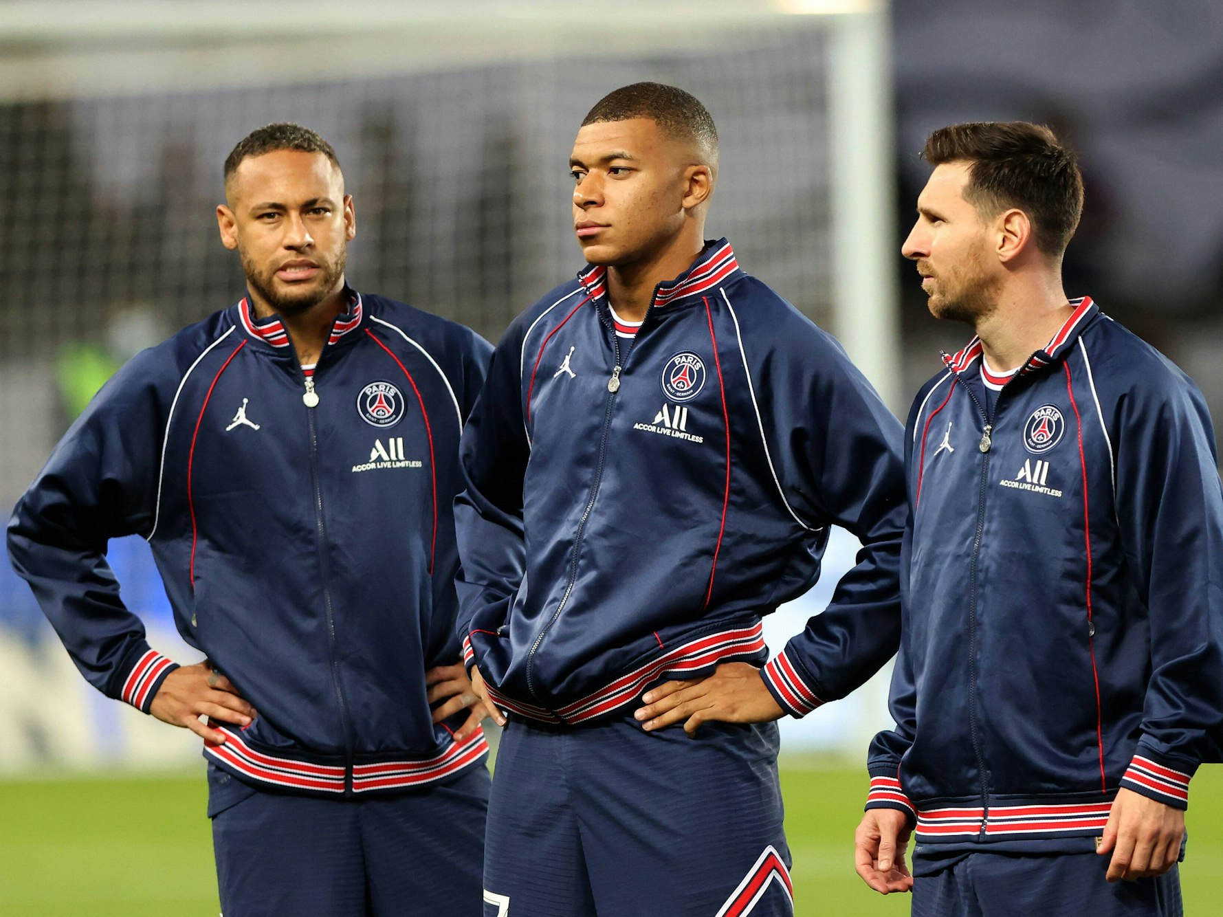 Neymar, Kylian Mbappe und Lionel Messi stehen in Trainingsjacken auf dem Feld. Neymar und Mbappe haben die Hände in den Hüften.