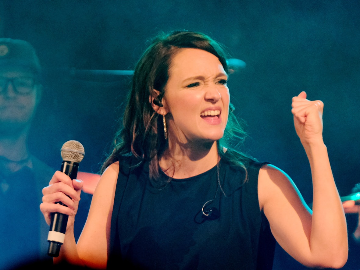 Carolina Kebekus steht mit einem Mikrofon in der Hand auf einer Bühne.