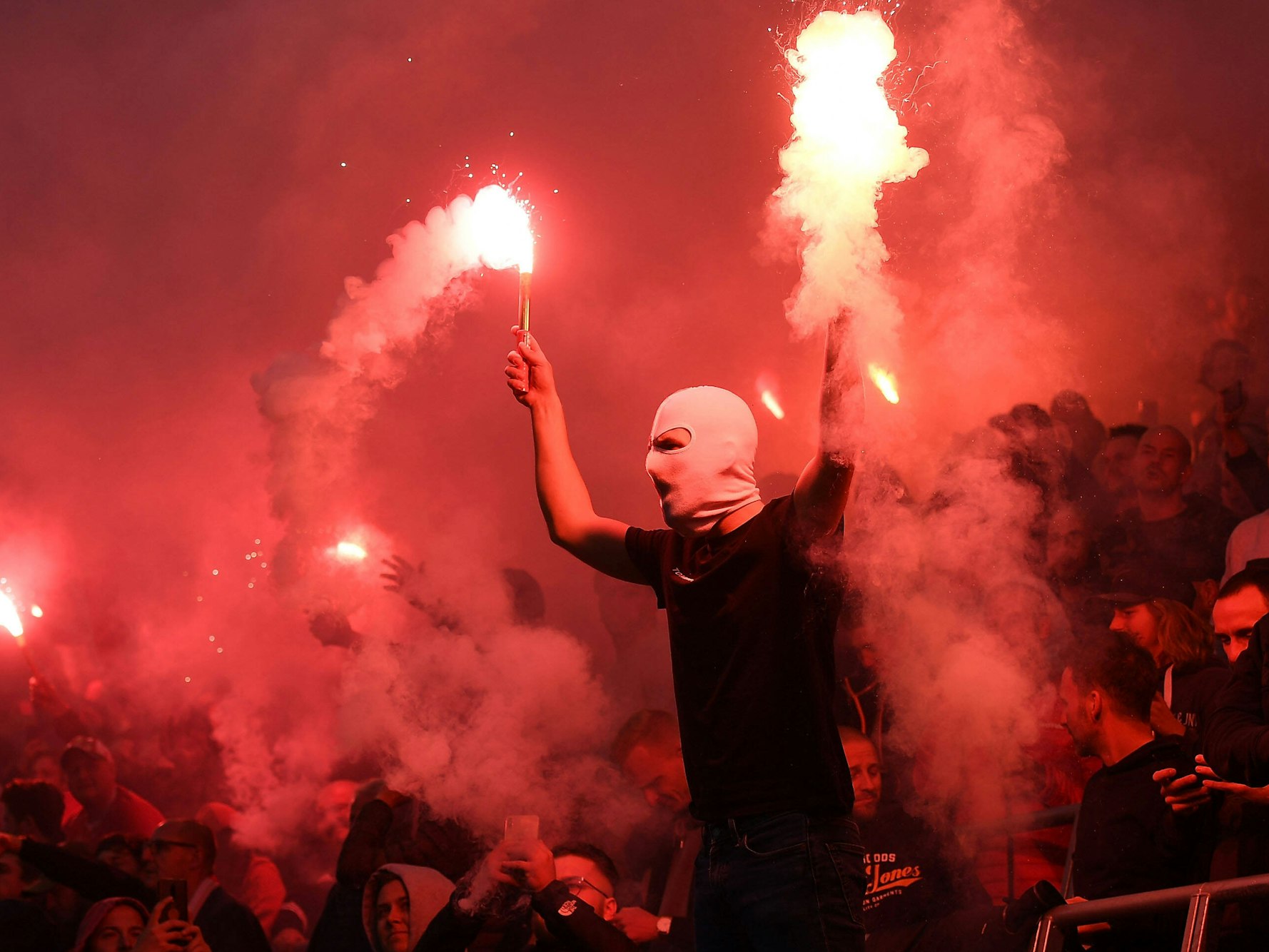 Ein vermummter steht auf einem Geländer und hält zwei Pyro-Fackeln in den Händen.