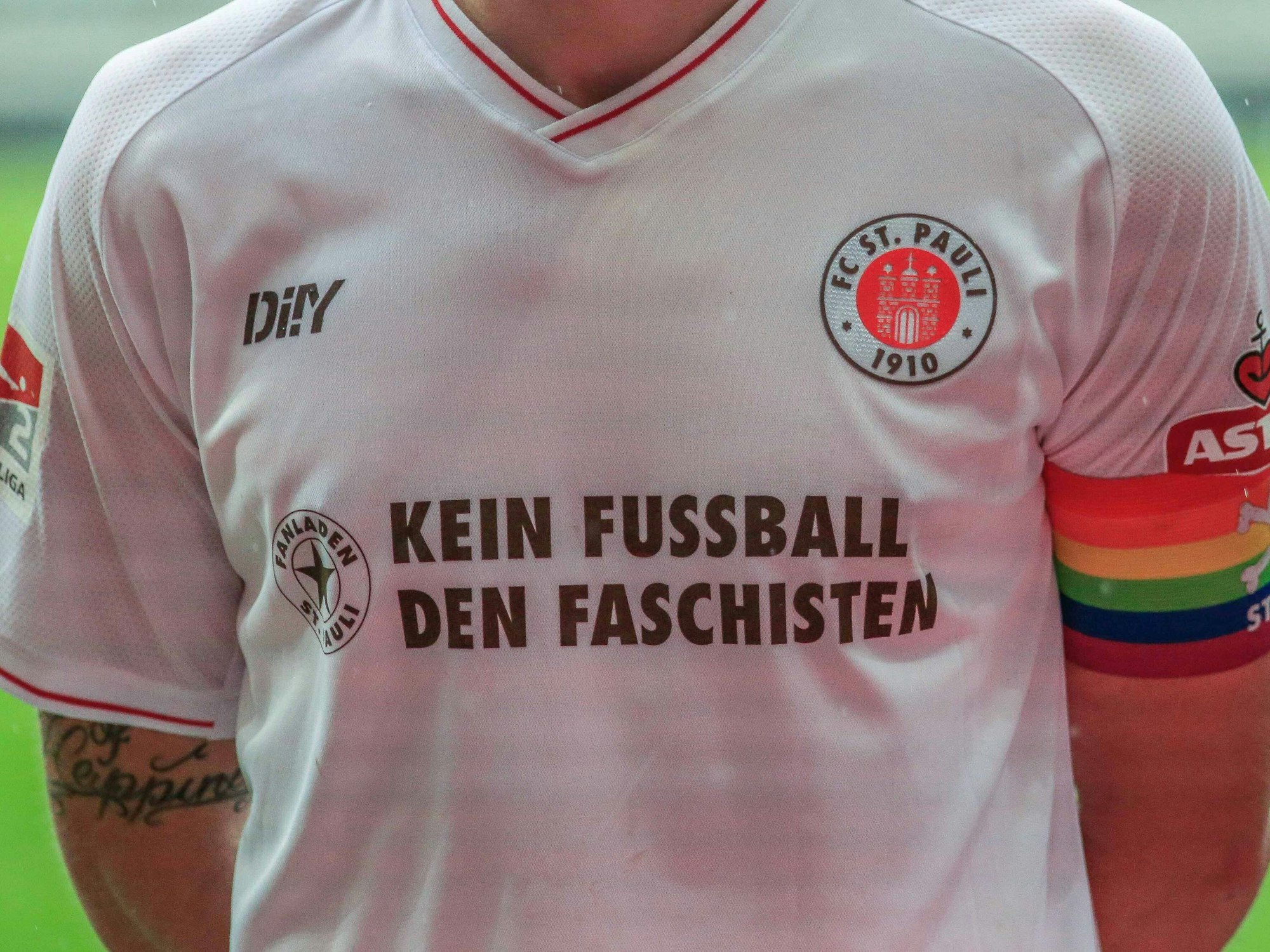 Auf der Brust des weißen Auswärtstrikots des FC St. Pauli ist „Kein Fußball den Faschisten“ zu lesen. Am Arm trägt die Person, deren Kopf nicht zu sehen ist, eine Binde in Regenbogenfarben.