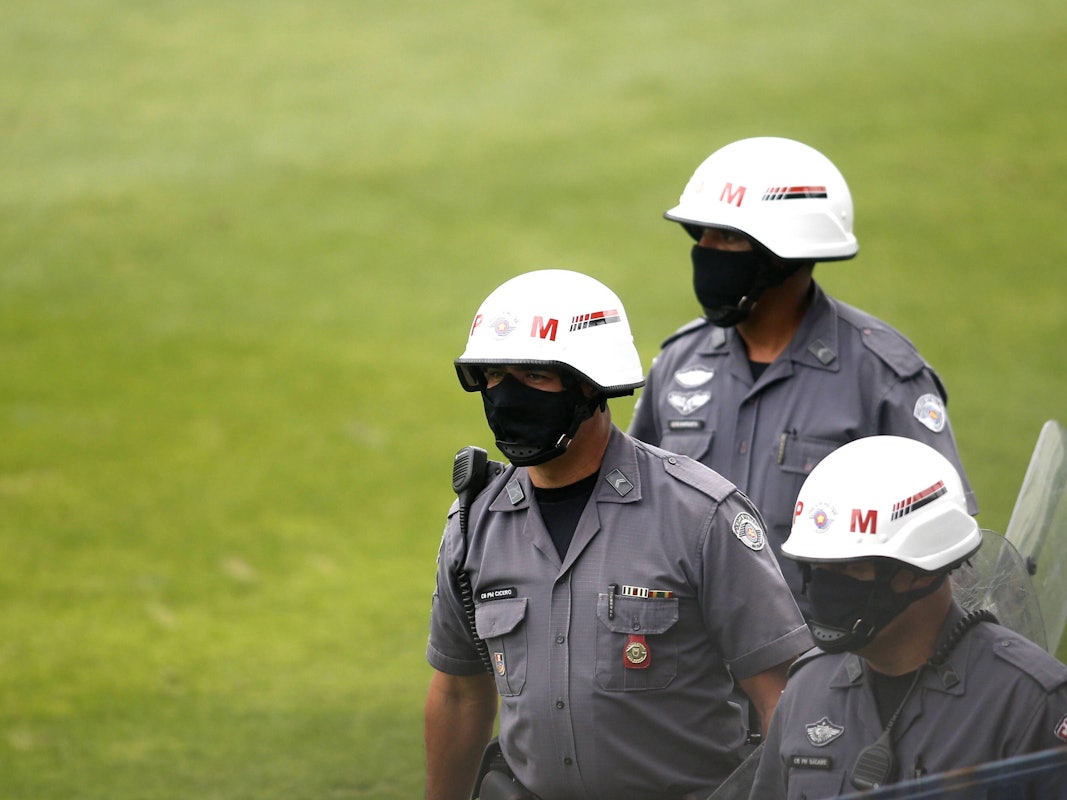 Drei uniformierte Militärpolizisten laufen über das Feld. Sie tragen Helme und Masken. Einer trägt ein Schild in der Hand.