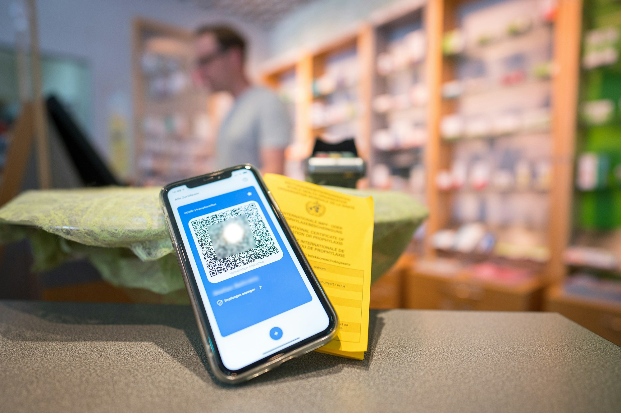 Ein Smartphone mit dem QR Code eines digitalen Impfausweises liegt zusammen mit einem Impfbuch auf dem Tisch einer Apotheke.