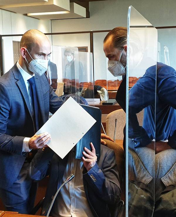 Prozessauftakt: Der Angeklagte hält sich Akten vor das Gesicht. Die Rechtsanwälte verdecken den Angeklagten mit Dokumenten.