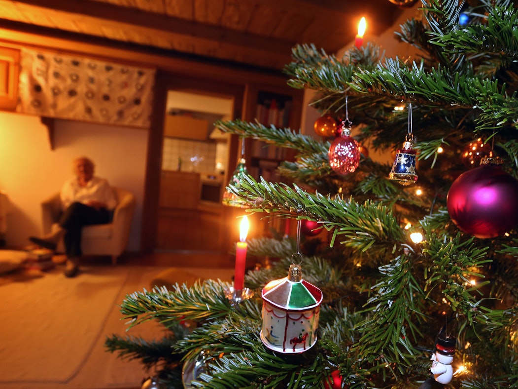 Auf dem Foto sieht man im rechten Teil des Bildes einen geschmückten Tannenbaum, im Hintergrund sitzt eine ältere Frau.