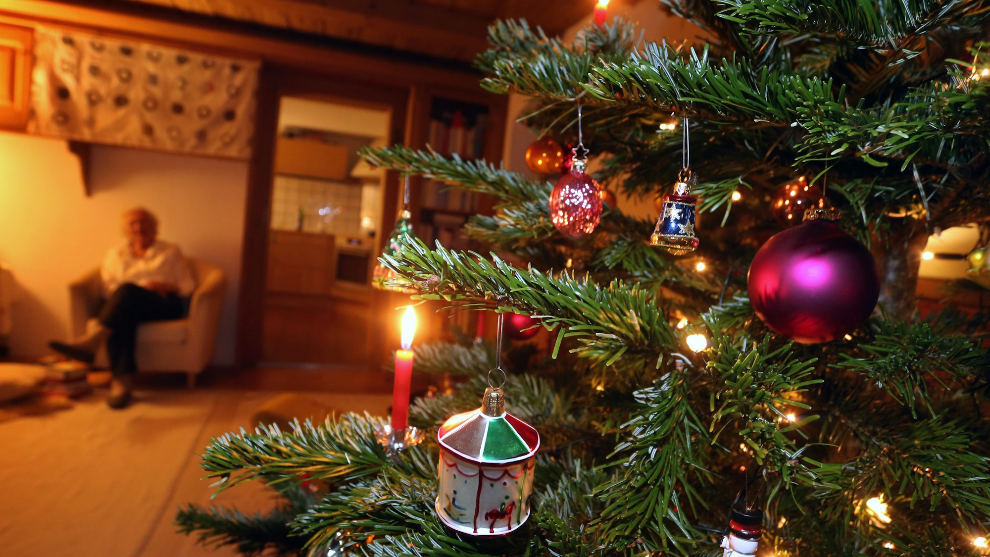 Auf dem Foto sieht man im rechten Teil des Bildes einen geschmückten Tannenbaum, im Hintergrund sitzt eine ältere Frau.