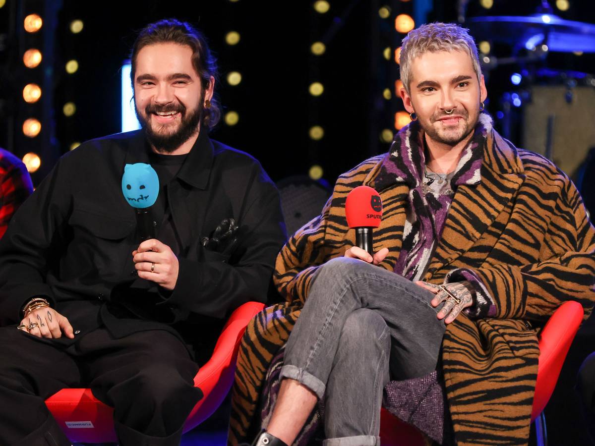 Tom und Bill Kaulitz von der Band Tokio Hotel sitzen während einer Radioshow auf einer Bühne mit jeweils einem Mikrofon in der Hand.