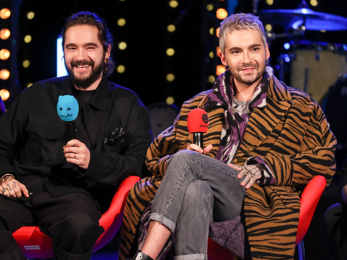Tom und Bill Kaulitz von der Band Tokio Hotel sitzen während einer Radioshow auf einer Bühne mit jeweils einem Mikrofon in der Hand.