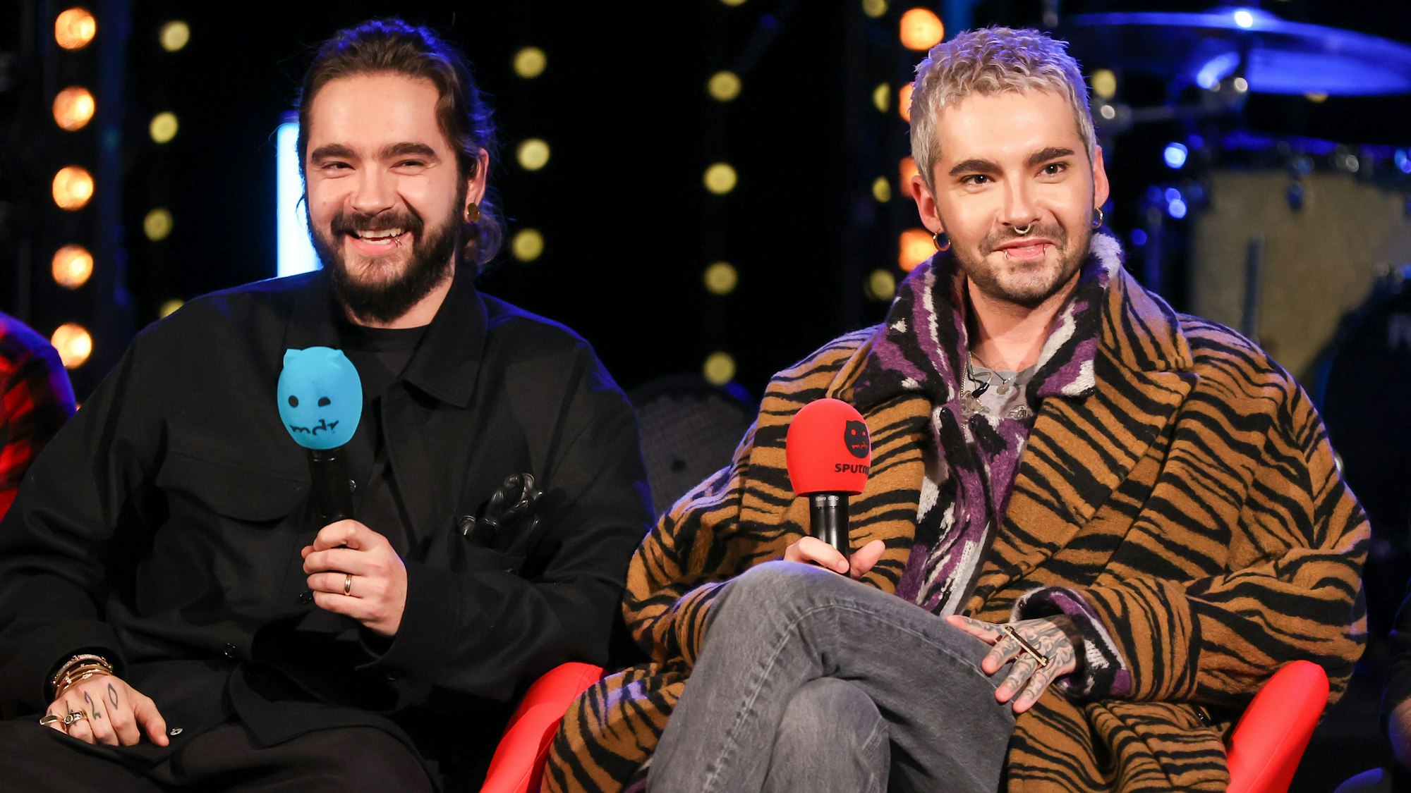 Tom und Bill Kaulitz von der Band Tokio Hotel sitzen während einer Radioshow auf einer Bühne mit jeweils einem Mikrofon in der Hand.