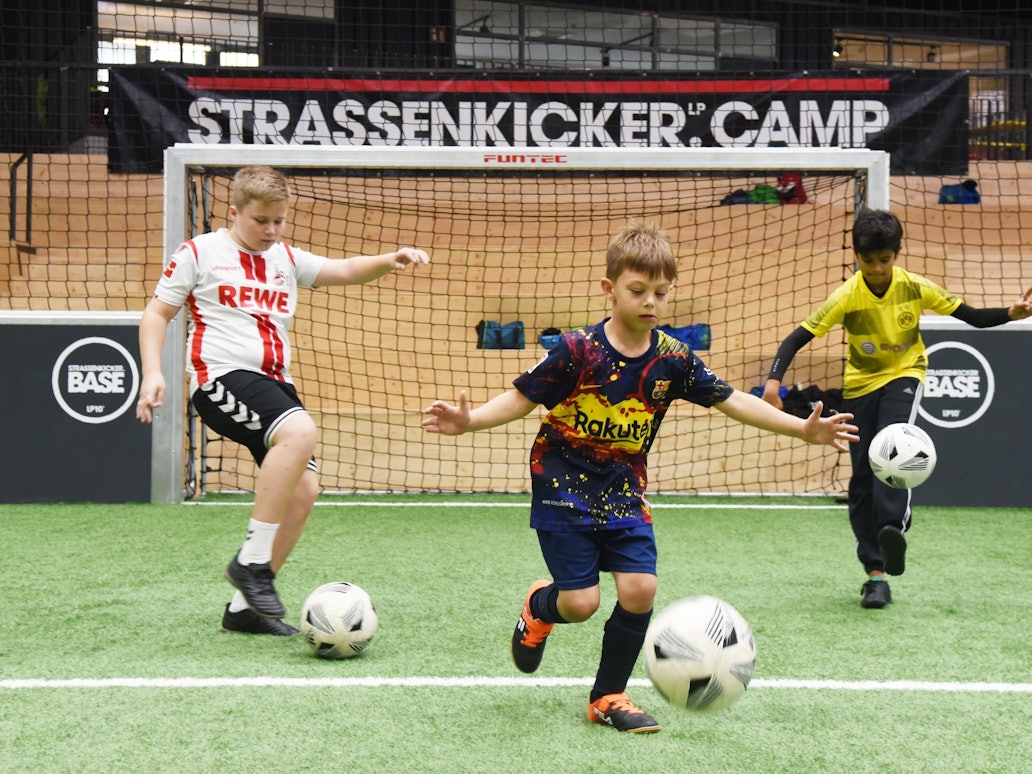 Drei Jungen (v.l Mats, Ilyas und Tom) dribbeln beim „Strassenkicker.Camp” in Köln-Mülheim auf einem Kunstrasenplatz.