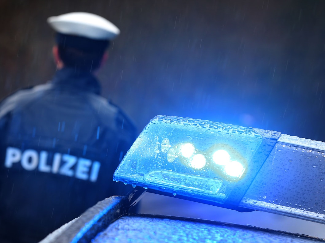 Ein Polizist steht im Regen vor einem Streifenwagen dessen Blaulicht aktiviert ist.
