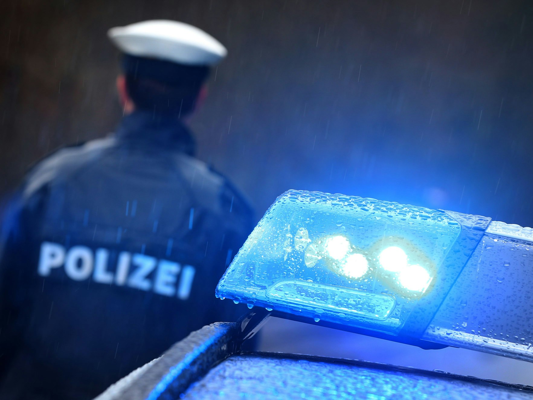 Ein Polizist steht im Regen vor einem Streifenwagen dessen Blaulicht aktiviert ist.
